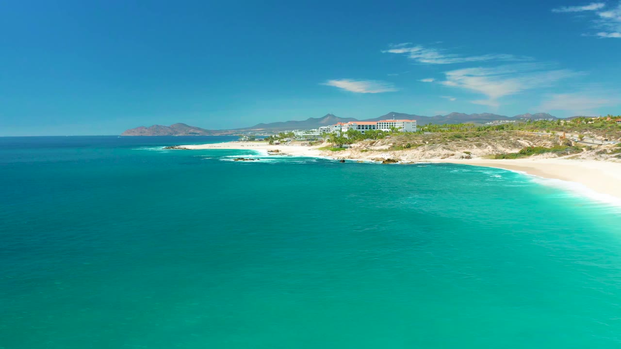 A second aerial view at Marquis Los Cabos, Mexico keeps the turquoise ocean broad in frame while the pale beach, low shoreline buildings, distant mountains, and clear sky remain visible with no visible people.