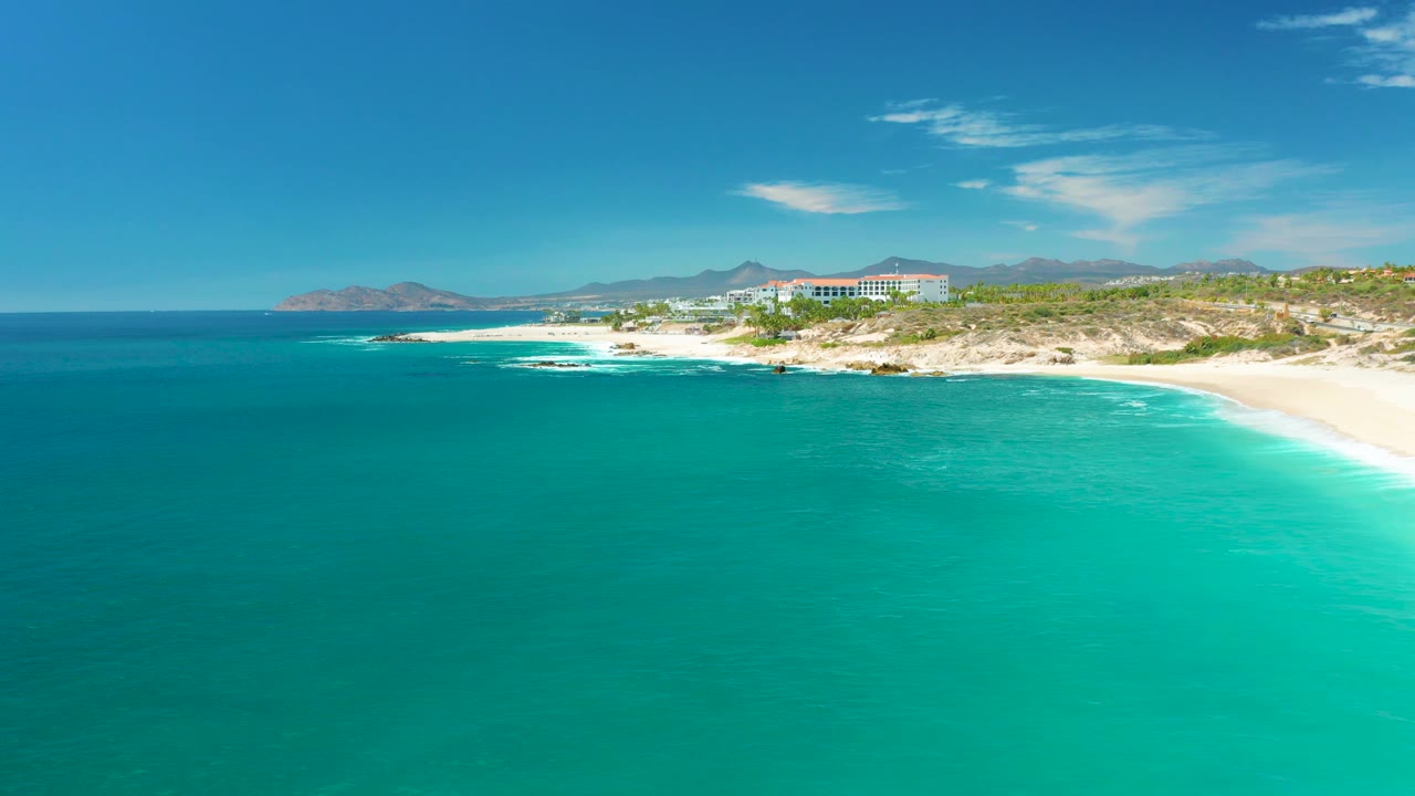 At Marquis Los Cabos, Mexico, this frame shows turquoise water meeting white surf near the beach, with rocky coastal edges, resort-area buildings, distant mountains, clear sky, and no visible people.