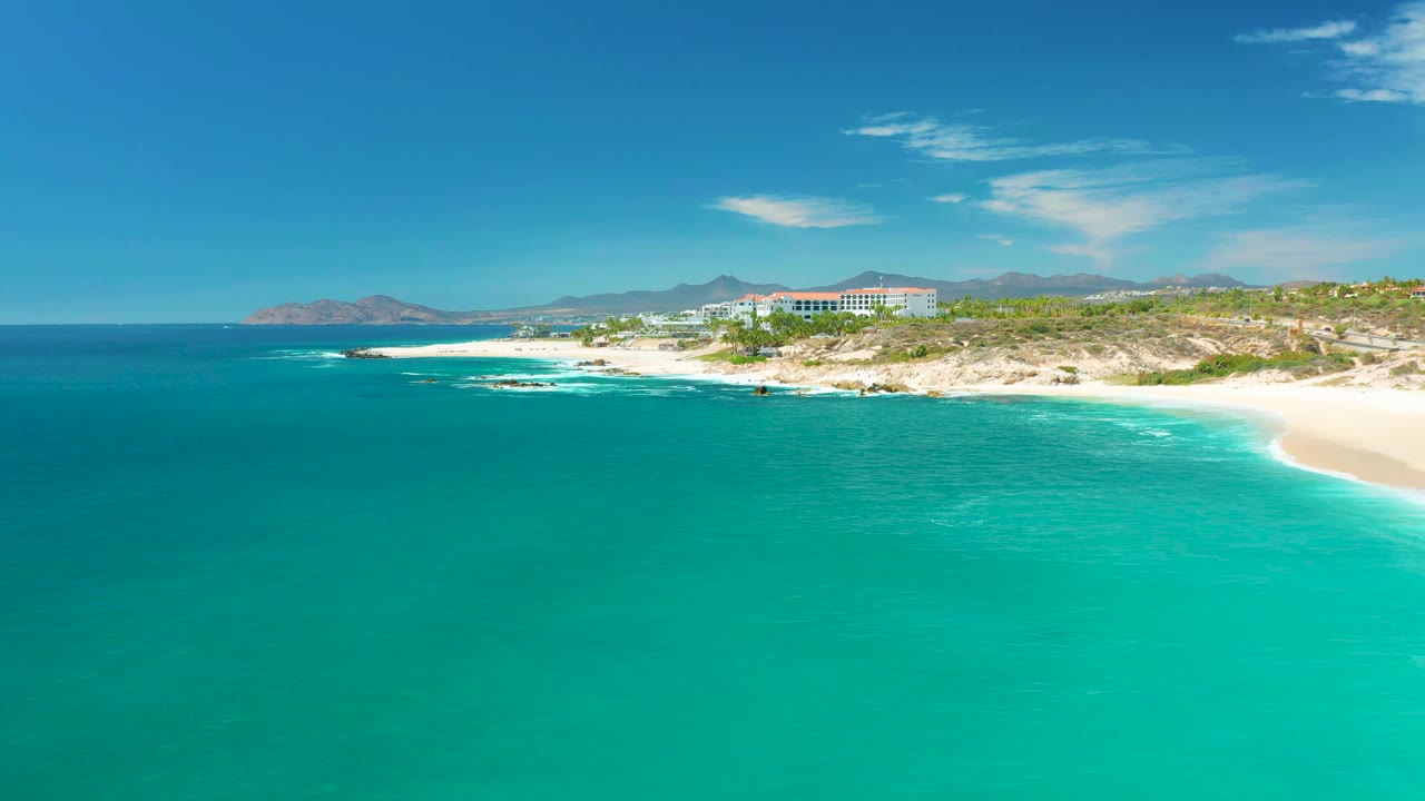 At Marquis Los Cabos, Mexico, the aerial view holds over turquoise water with thin white surf near the shore, a sandy beach at right, coastal buildings above the rocks, and no visible people.