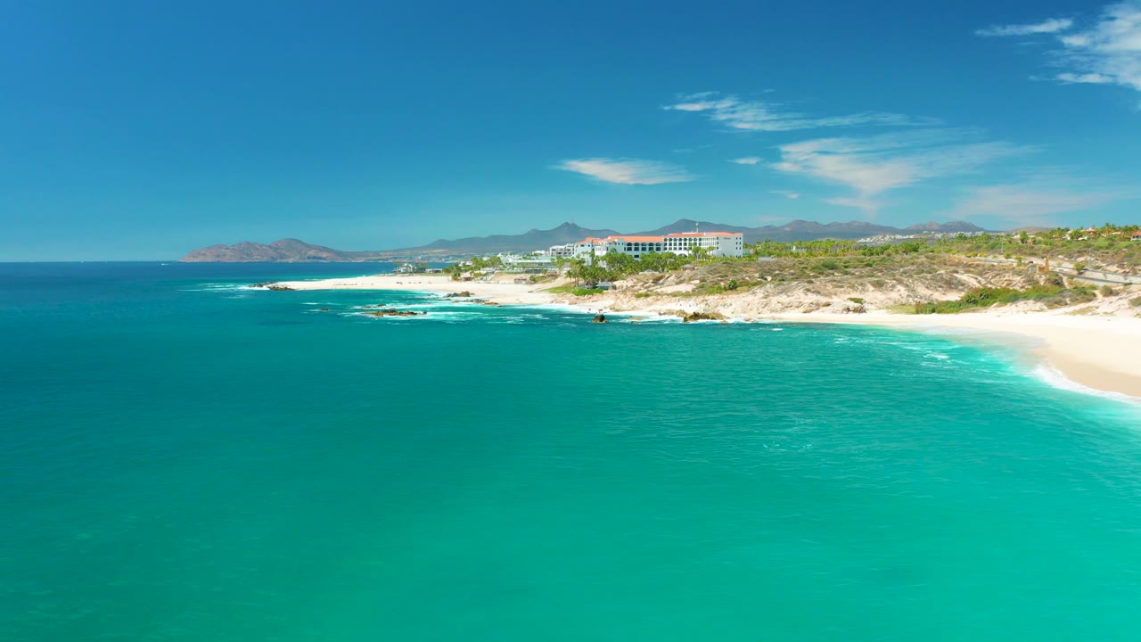 At Marquis Los Cabos, Mexico, open turquoise water dominates the foreground, white waves break along the rocky coast, the sandy beach stays at the right edge, and no visible people are present.