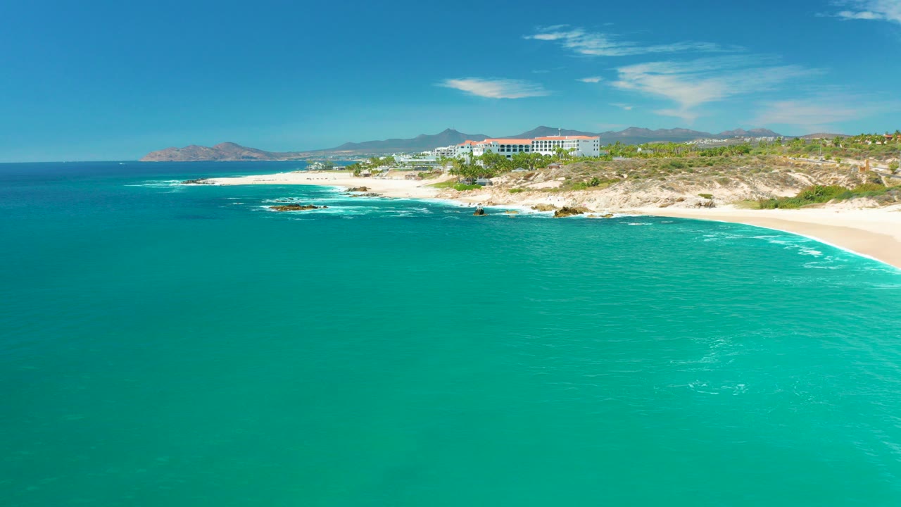 Aerial view at Marquis Los Cabos showing turquoise ocean in the foreground, white surf along the sandy and rocky shore, coastal buildings on the hillside, mountains in back, and no visible people.