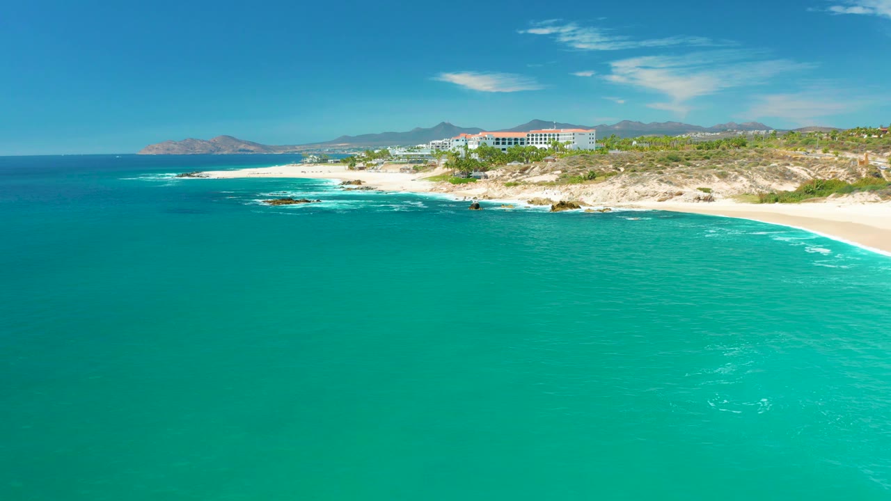 Marquis Los Cabos coastline seen from above, with blue-green ocean across the foreground, foamy surf at the shore, hillside vegetation and buildings, mountains beyond, and no visible people.