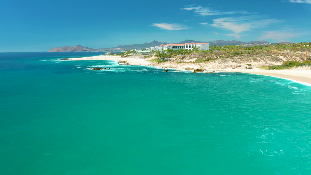 Aerial view of the Marquis Los Cabos shore with clear turquoise water in front, whitecaps near rocks and sand, the coastal building cluster on the hillside, distant mountains, and no visible people.
