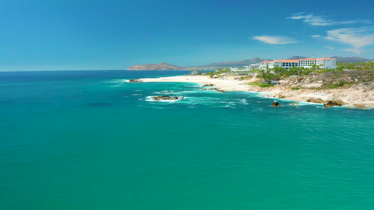 Wide aerial view of the Marquis Los Cabos shoreline with turquoise water in the foreground, sandy beach and rocky outcrops near the center, coastal buildings at upper right, and no visible people.