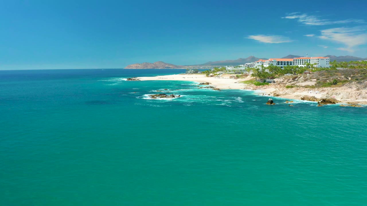 Aerial frame at Marquis Los Cabos showing turquoise ocean across most of the view, white waves around dark rocks, a pale sandy beach, low coastal buildings, and no visible people.