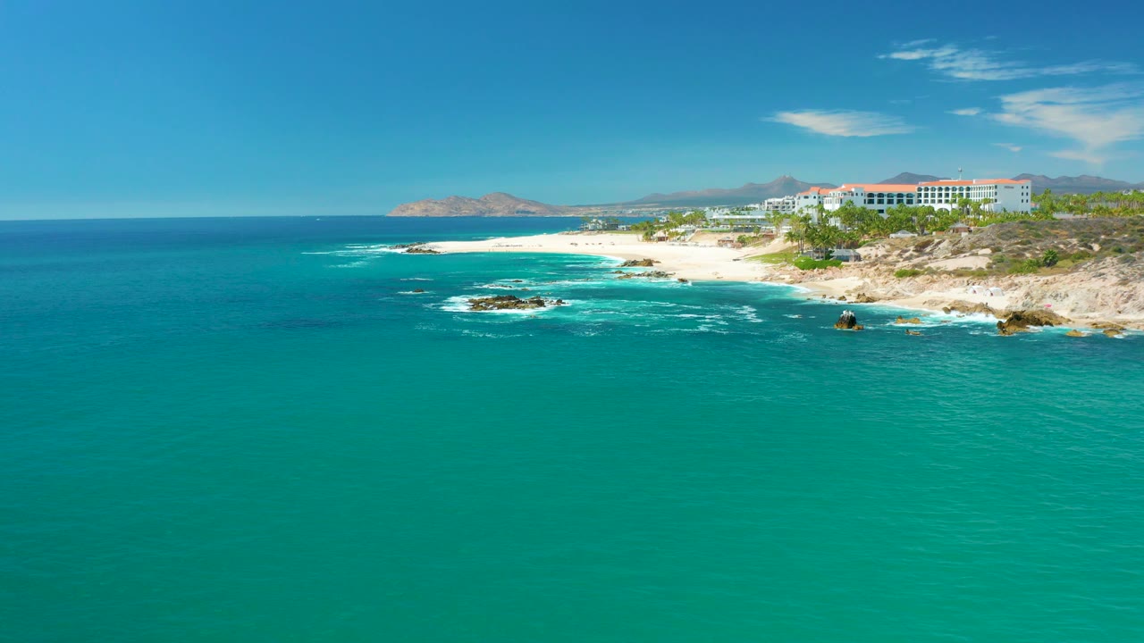 Aerial coastline with turquoise ocean, white surf, rocks, beach, and buildings.