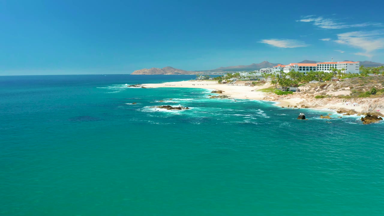 At Marquis Los Cabos, Mexico, white foam spreads around offshore rocks beside the sandy beach as coastal buildings remain visible in the background and no people are visible.
