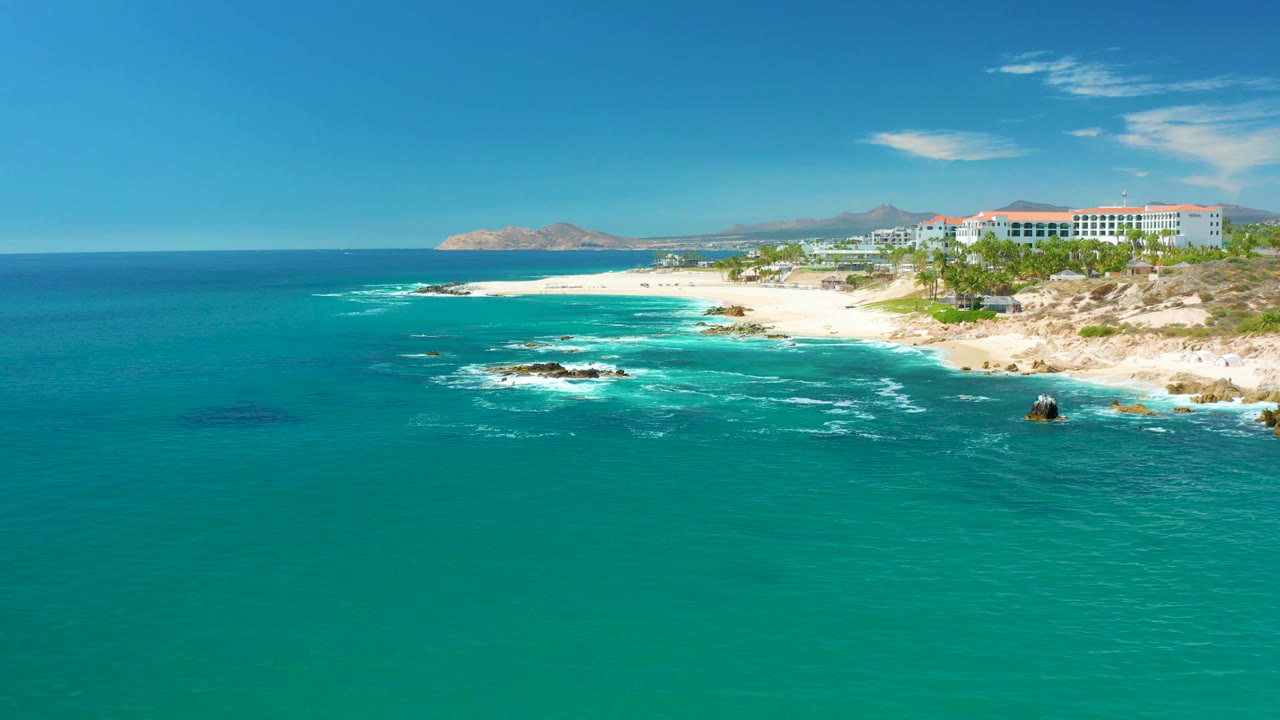 At Marquis Los Cabos, Mexico, waves break in a loose white arc over rocks near the sandy beach, with turquoise water in front and coastal buildings beyond, with no visible people.