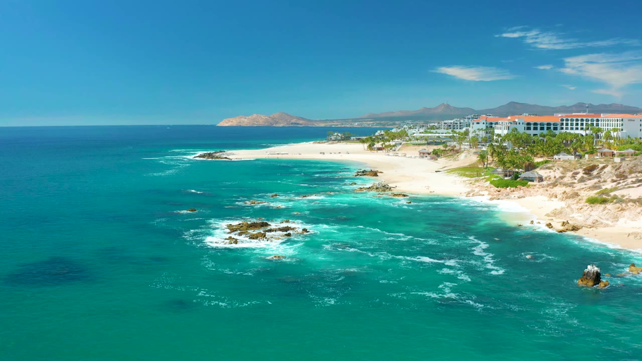 Aerial coastal frame from Marquis Los Cabos showing white foam widening over shallow turquoise water, rocks in the surf, the sandy shoreline to the right, and no visible people.