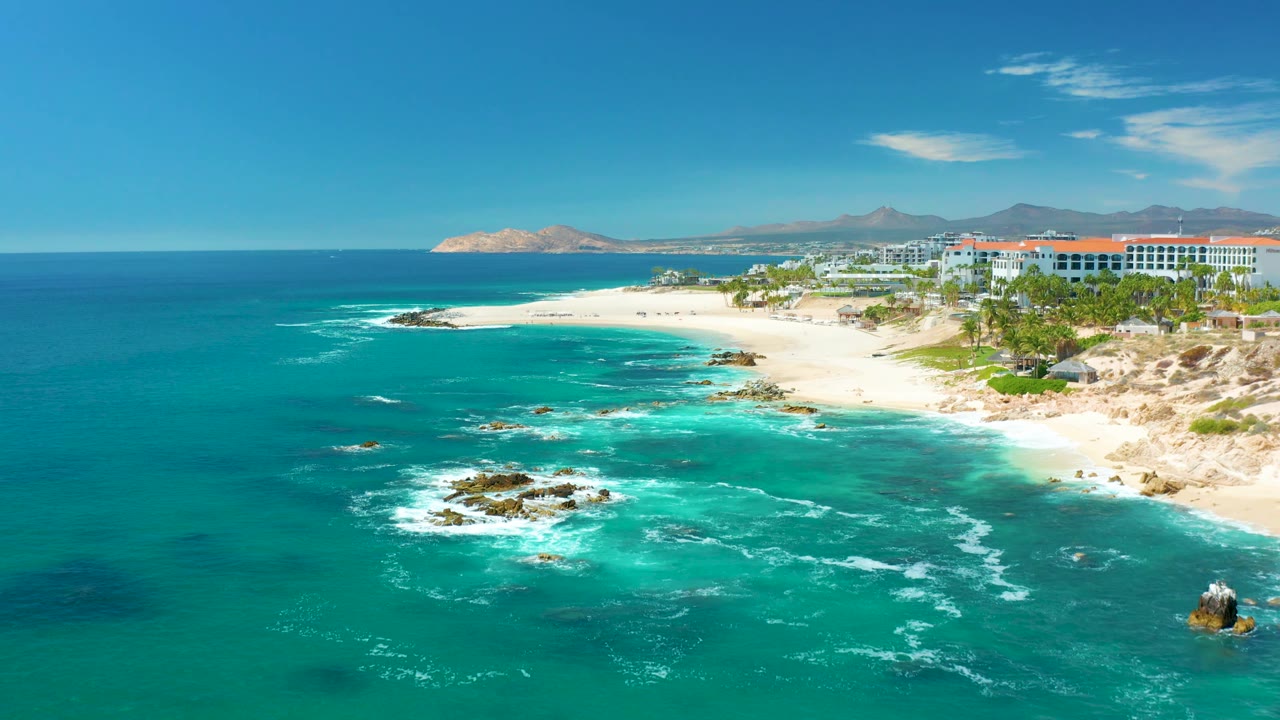Wide aerial coastline with surf, rocks, sand, buildings, and blue sky.