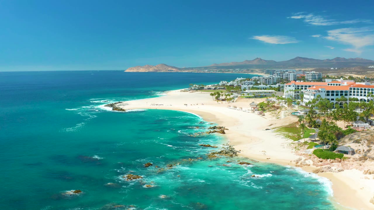 Aerial view at Marquis Los Cabos, Mexico with deeper blue water offshore, turquoise shallows near rocks, a curved sandy shore, coastal buildings, and no visible people.