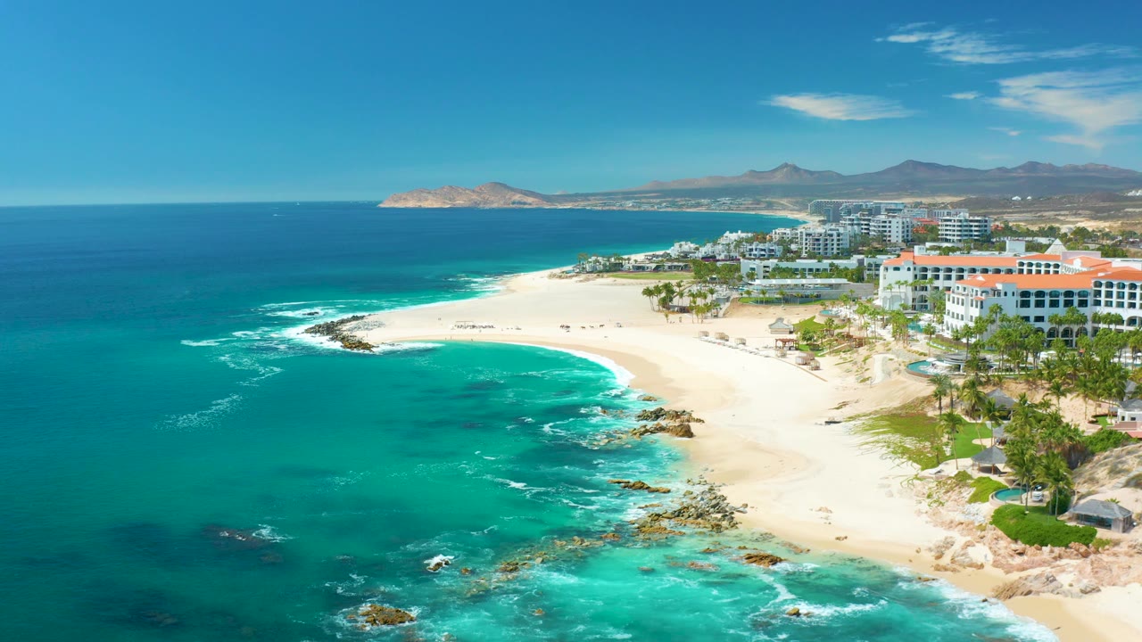 Aerial frame from Marquis Los Cabos, Mexico with a wider sweep of open ocean on the left, a bright sandy point, shoreline rocks, buildings and palms on the right, and no visible people.