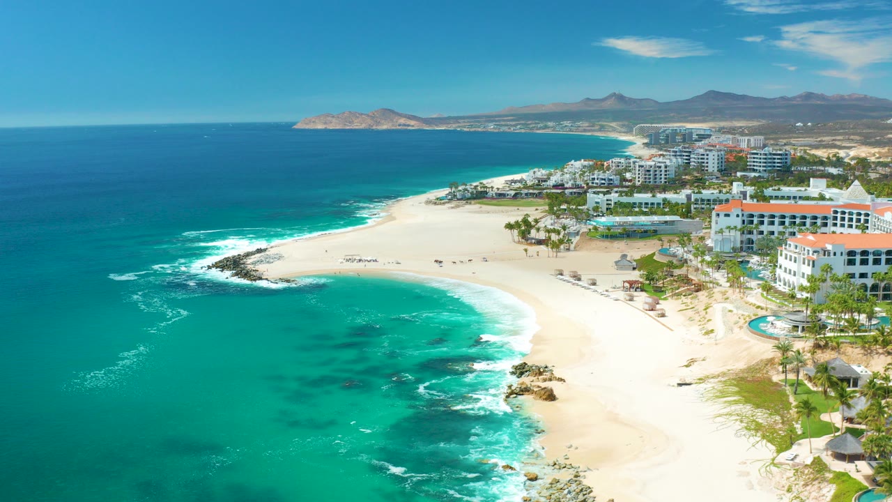 Aerial frame over Marquis Los Cabos, Mexico with turquoise water in the foreground, white surf on pale sand, resort buildings at right, and distant mountains under a clear sky.