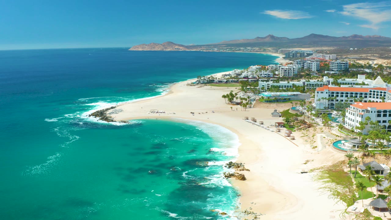 The frame holds a high view over Marquis Los Cabos with deeper blue water offshore, turquoise shallows, pale beach, resort structures to the right, and mountains beyond the coast.