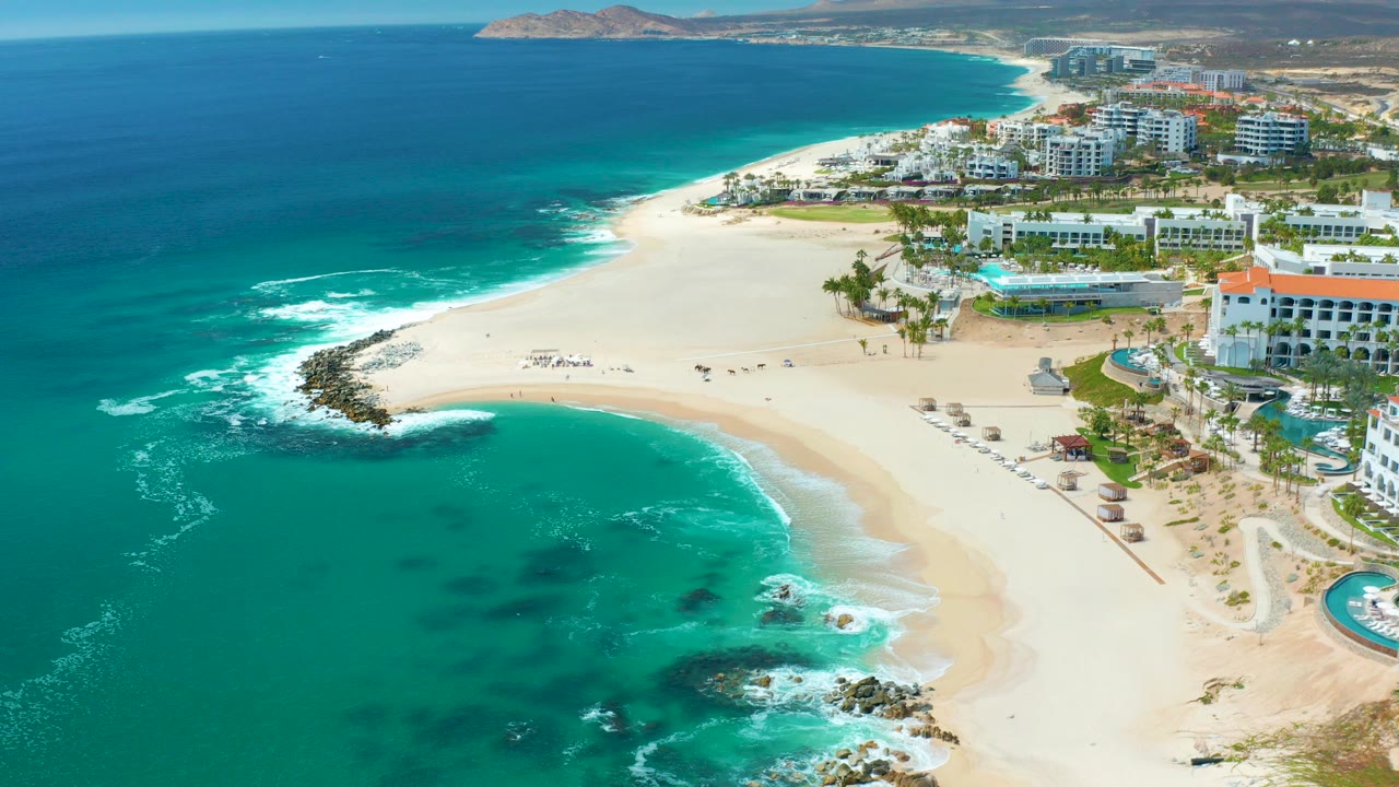 Aerial frame over Marquis Los Cabos, Mexico showing turquoise water around dark rocks, a broad sandy beach, small beach setups, scattered people, and white coastal buildings beyond the shore.