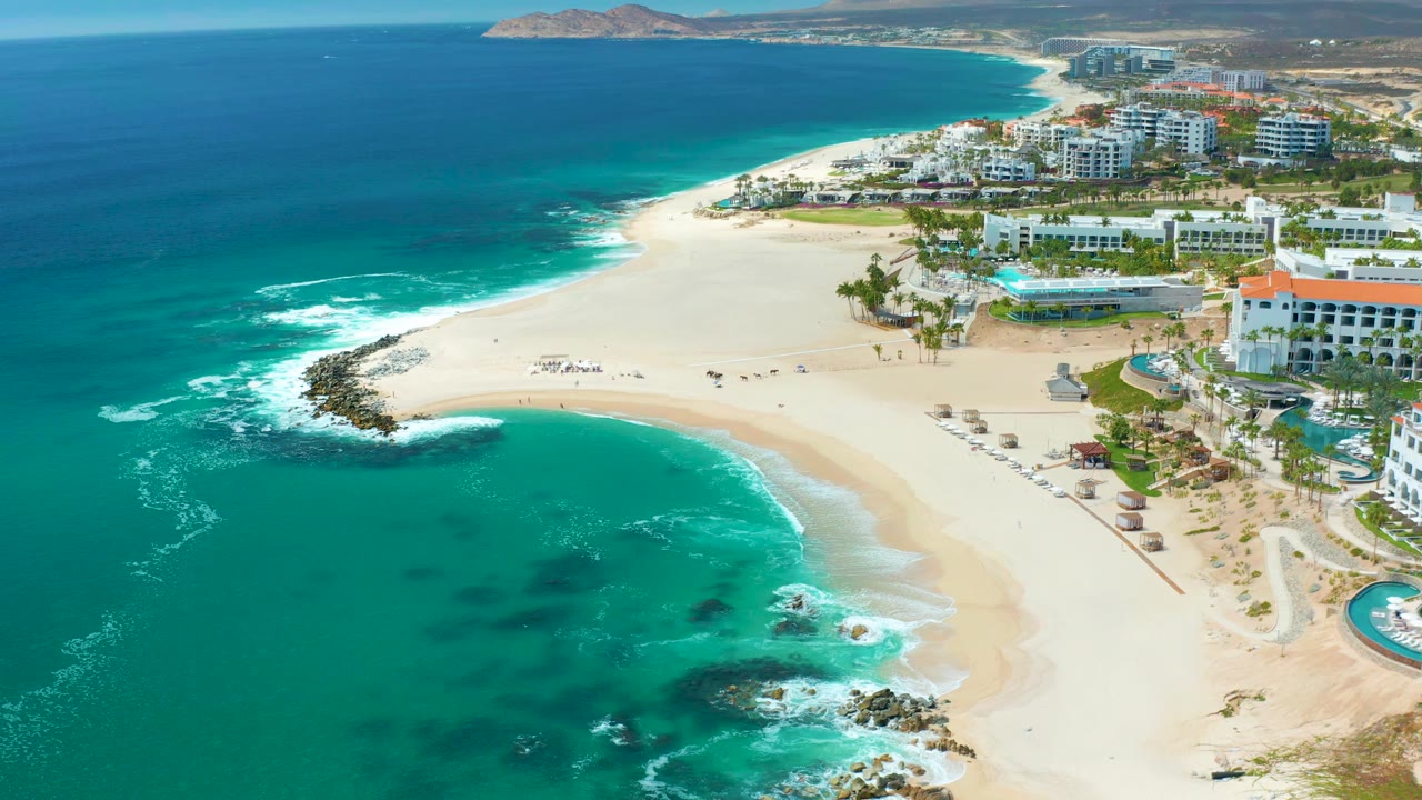 Drone view of a curving sandy shoreline with waves and resort buildings.