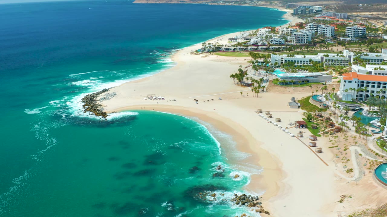 High aerial frame over Marquis Los Cabos, Mexico showing blue-green ocean on the left, pale beach on the right, a dark rock point, umbrella rows, visible beachgoers, and hotel buildings behind the sand.