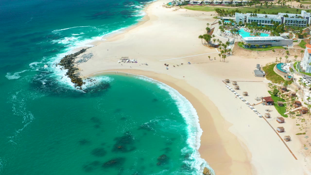 Aerial frame over Marquis Los Cabos in Mexico showing turquoise water in the foreground, white surf on a curved sandy beach, dark shoreline rocks, umbrellas, pools, and resort buildings at the top edge.