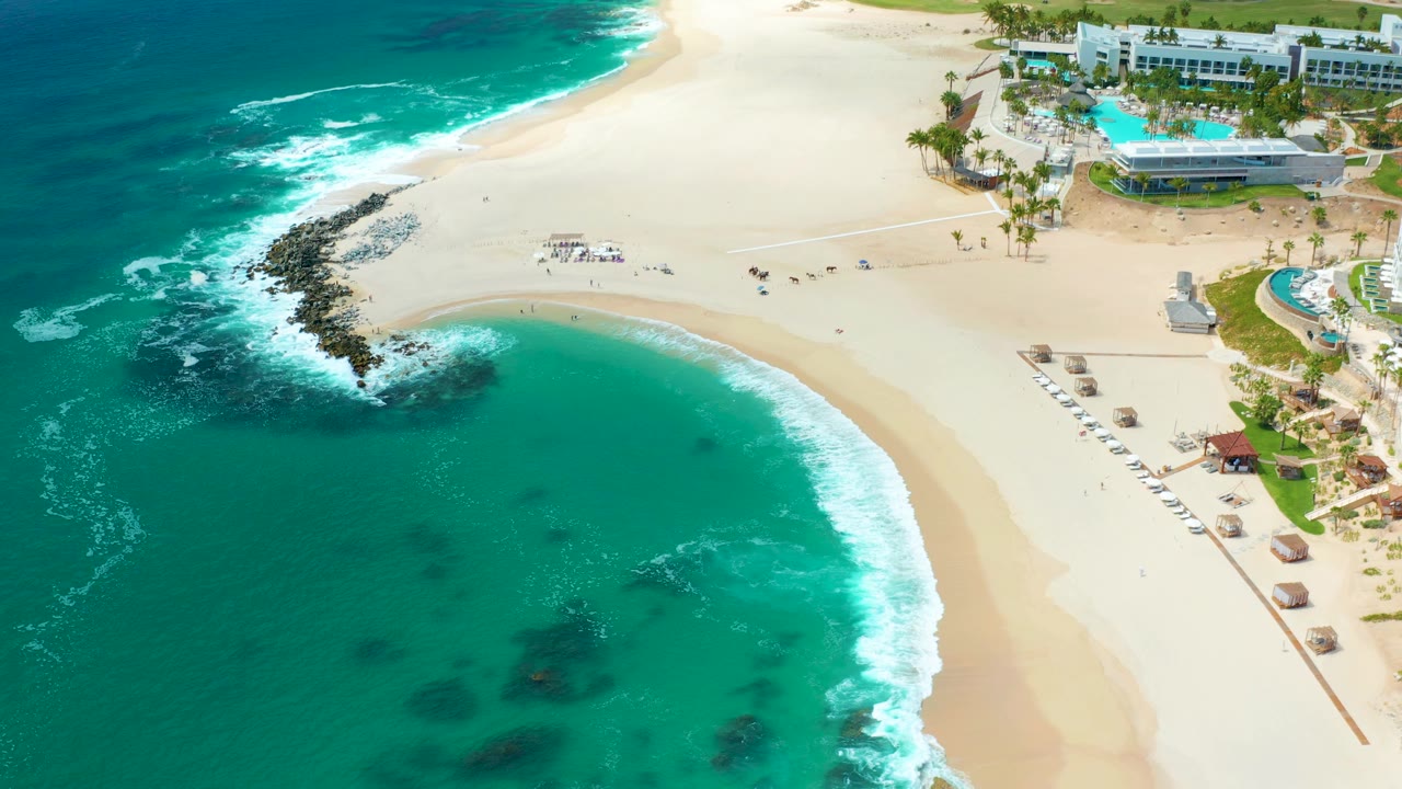 High drone frame over Marquis Los Cabos showing stronger white water along the upper shoreline, a rocky outcrop left of center, pale sand with scattered small people, and resort buildings and pools along the top right.