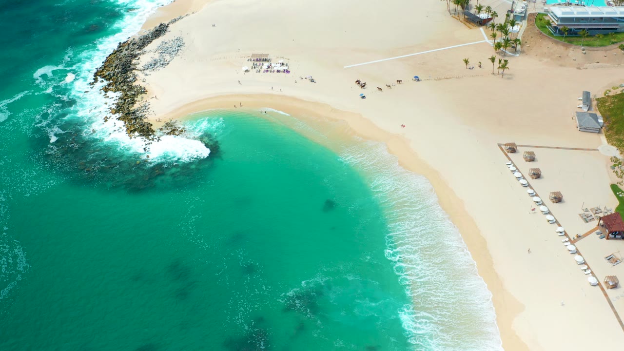 High aerial of clear water and curved beach with surf breaking on rocks.