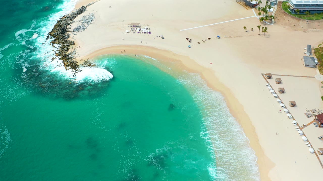 The cove remains centered from above, with white foam spreading from the rocky point into turquoise water and scattered beachgoers visible on the sand at Marquis Los Cabos.