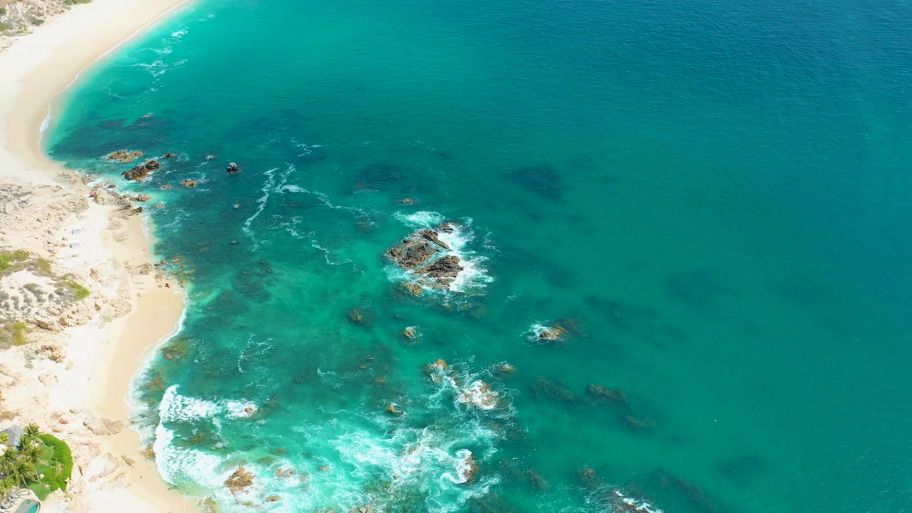 Aerial frame over Marquis Los Cabos, Mexico with the beach curving along the left edge, darker submerged rocks, foamy wave breaks, and no visible people.