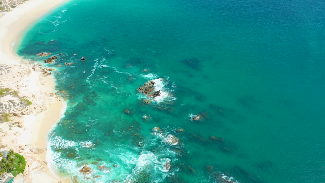 Aerial frame from Marquis Los Cabos, Mexico showing pale sand, bright turquoise shallows, dark reefs, a larger white wave patch near the lower left, and no visible people.