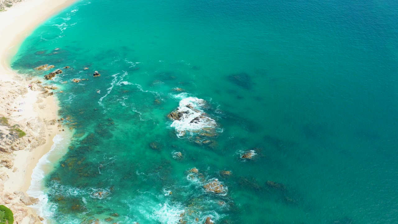 Aerial view of turquoise ocean, sandy beach, rocks, and white surf.