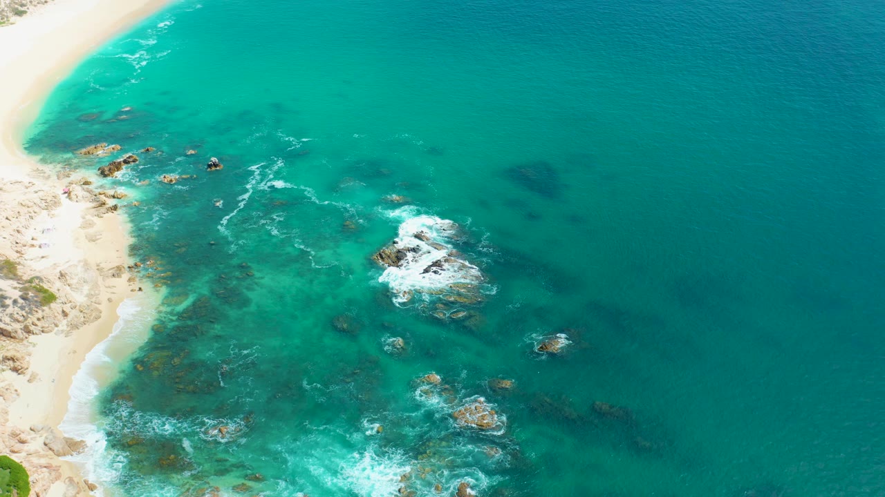 Aerial view at Marquis Los Cabos with the shoreline still along the left edge, darker rock patches under turquoise water, fresh white foam near the central rocks, and no visible people.