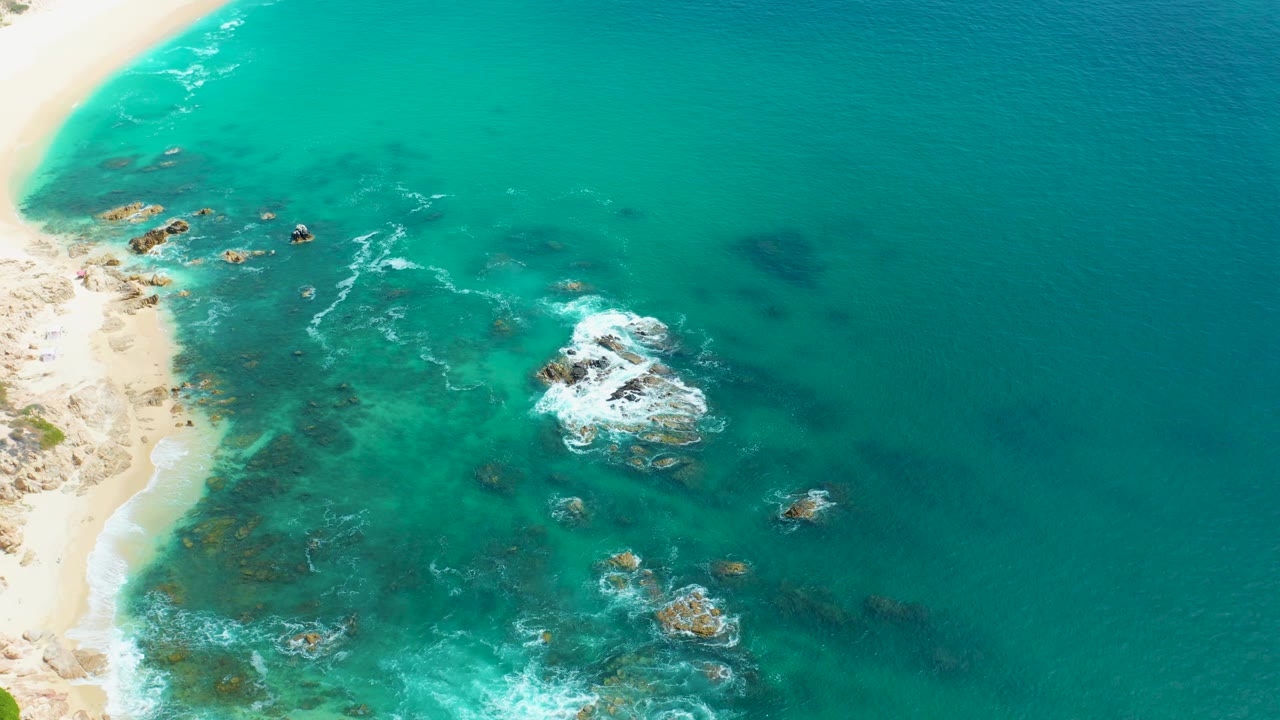 The Marquis Los Cabos coast appears from above with a narrow strip of sand at left, blue-green water across the frame, rock clusters near center, active foam, and no visible people.