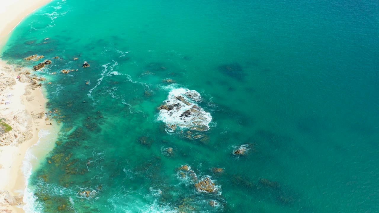 A high aerial frame at Marquis Los Cabos shows the sandy beach pushed to the left, a large foaming rock group near center right, smaller rocks below, and no visible people.