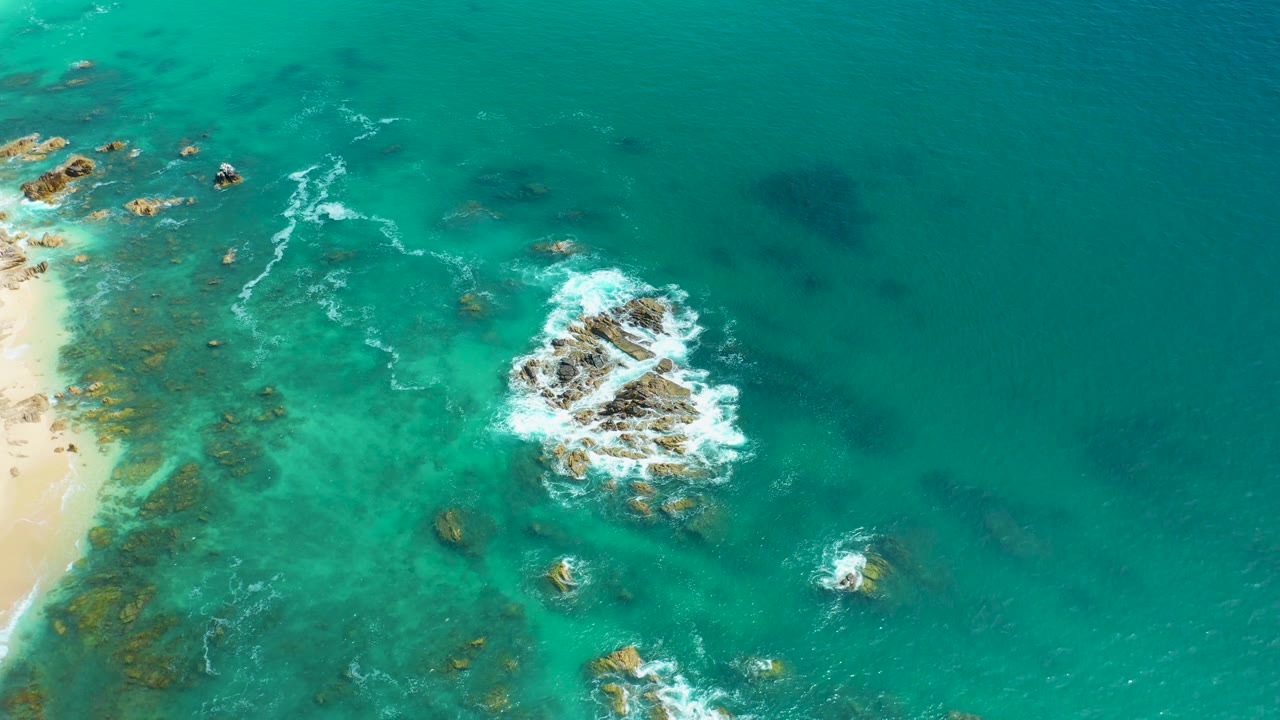 Aerial frame at Marquis Los Cabos, Mexico with the main rock formation near center, white foam spreading on its upper side, smaller rocks below, a thin beach edge at left, and no visible people.