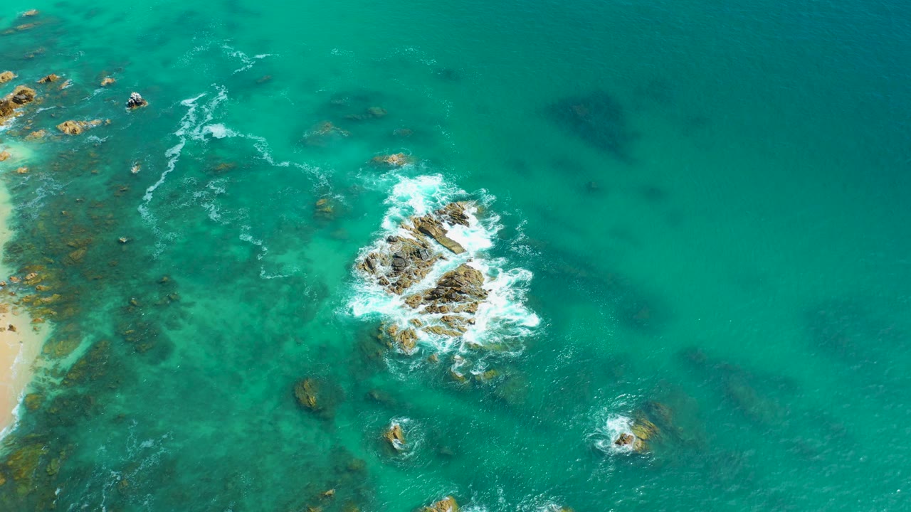 Top-down view at Marquis Los Cabos, Mexico with a larger central rock cluster, white foam curling around it, pale sand just inside the left border, and no visible people.
