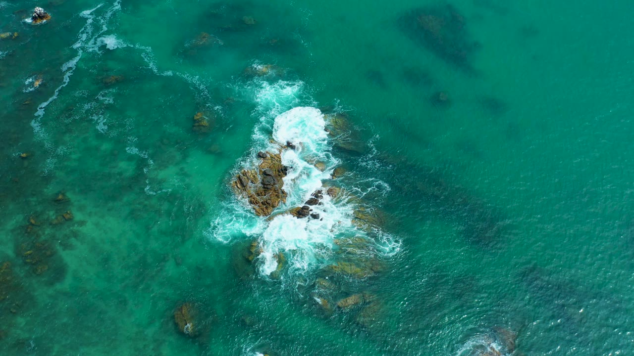 Aerial view at Marquis Los Cabos, Mexico with white surf expanding over the central rocks and darker submerged patches visible in the surrounding green-blue water, with no visible people.