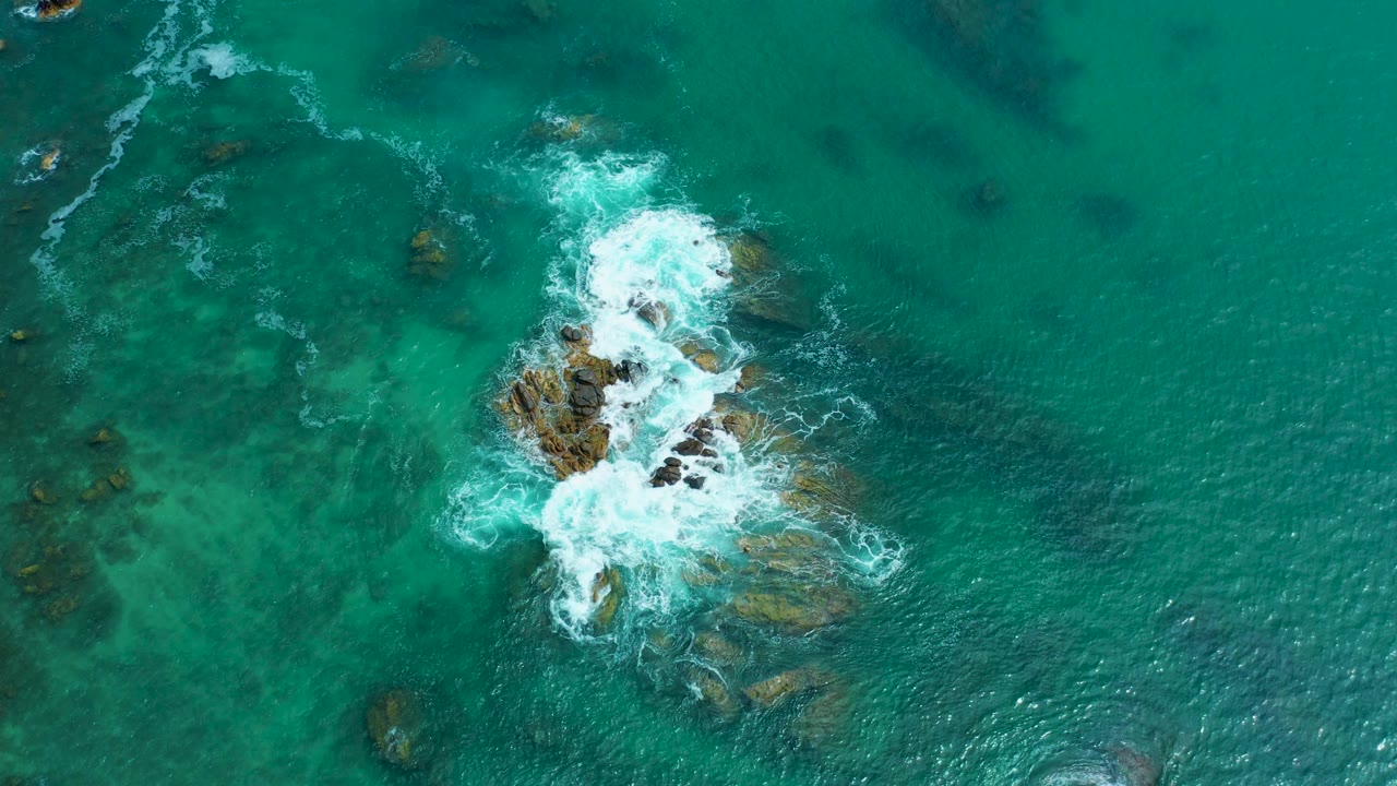 Top-down drone frame at Marquis Los Cabos, Mexico showing a larger burst of white foam wrapping the rocky seabed while open turquoise water fills the frame, with no visible people.