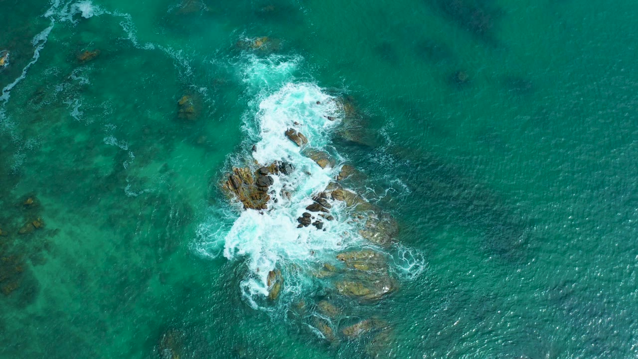 Aerial frame at Marquis Los Cabos, Mexico with the rocky seabed shifted lower in the composition as white foam pools on the left and upper edges, with no visible people.