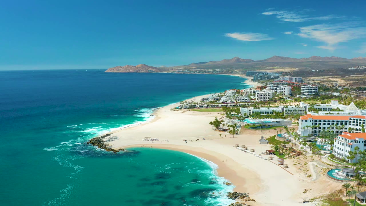 Wide aerial view at Marquis Los Cabos in Mexico with turquoise ocean on the left, pale sand curving around the shore, resort buildings to the right, distant people on the beach, and hills on the horizon.