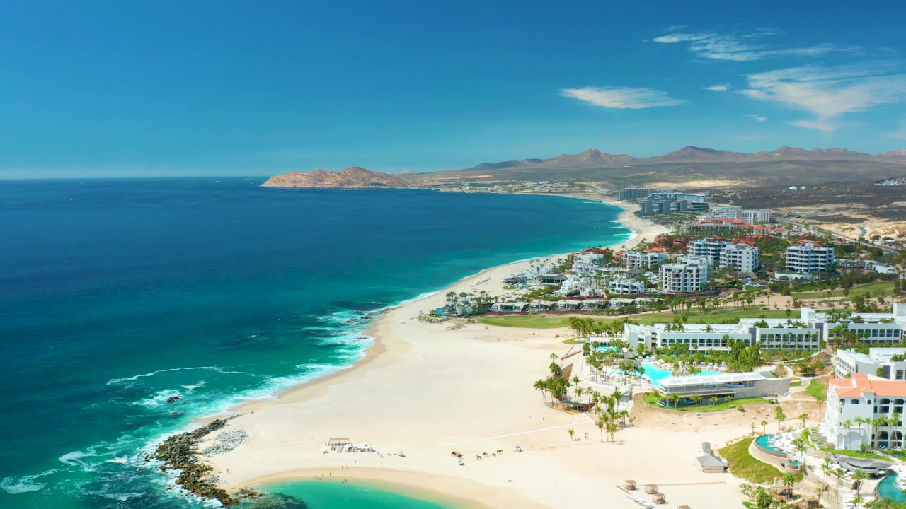 Aerial frame over Marquis Los Cabos showing the shoreline curve, turquoise water near the sand, white resort blocks, pools, palms, distant hills, and no visible people.