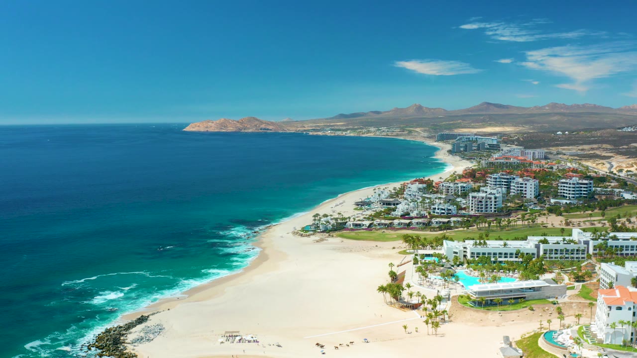 Mid-clip aerial view of Marquis Los Cabos with foam lines on the water, a bright sand beach, clustered resort buildings, pool areas, distant mountains, and no visible people.