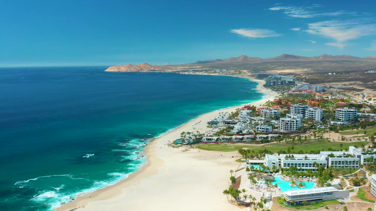 Aerial frame at Marquis Los Cabos, Mexico with the shoreline curving through the center, white waves on the sand, resort buildings on the right, mountains behind, and no visible people.