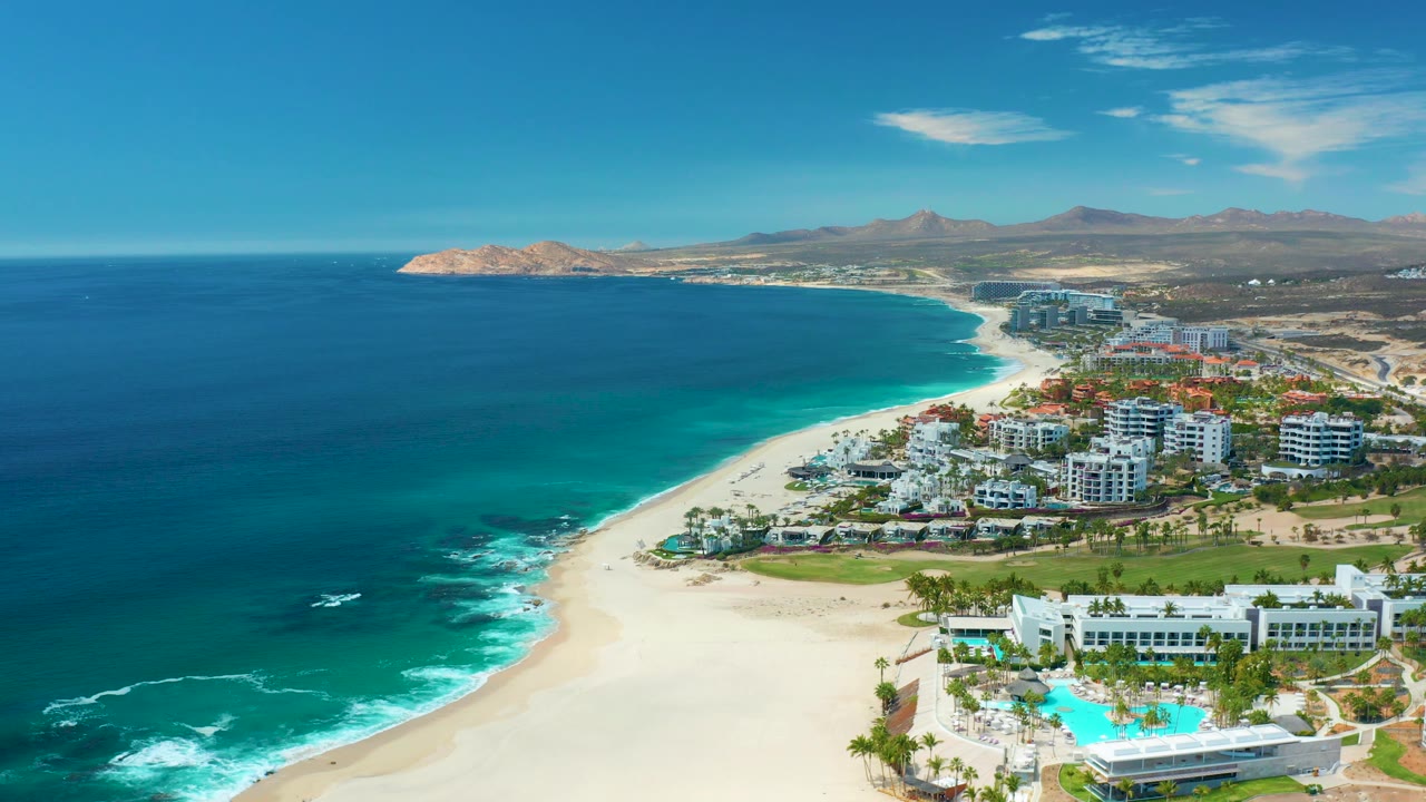 Aerial view of Marquis Los Cabos, Mexico showing turquoise shallows beside pale sand, small white surf breaks, resort buildings and pool area at right, dry mountains in back, and no visible people.