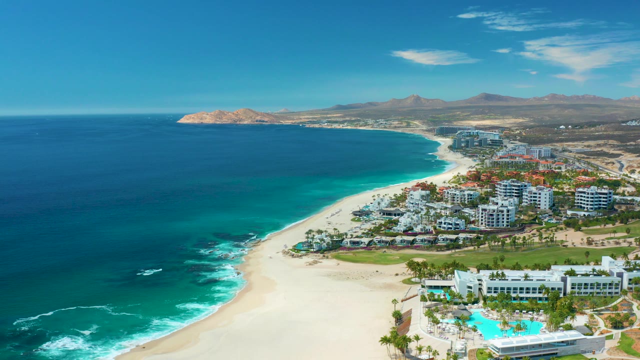Aerial coastline with blue water, surf on sand, resort buildings, pool, and mountains.
