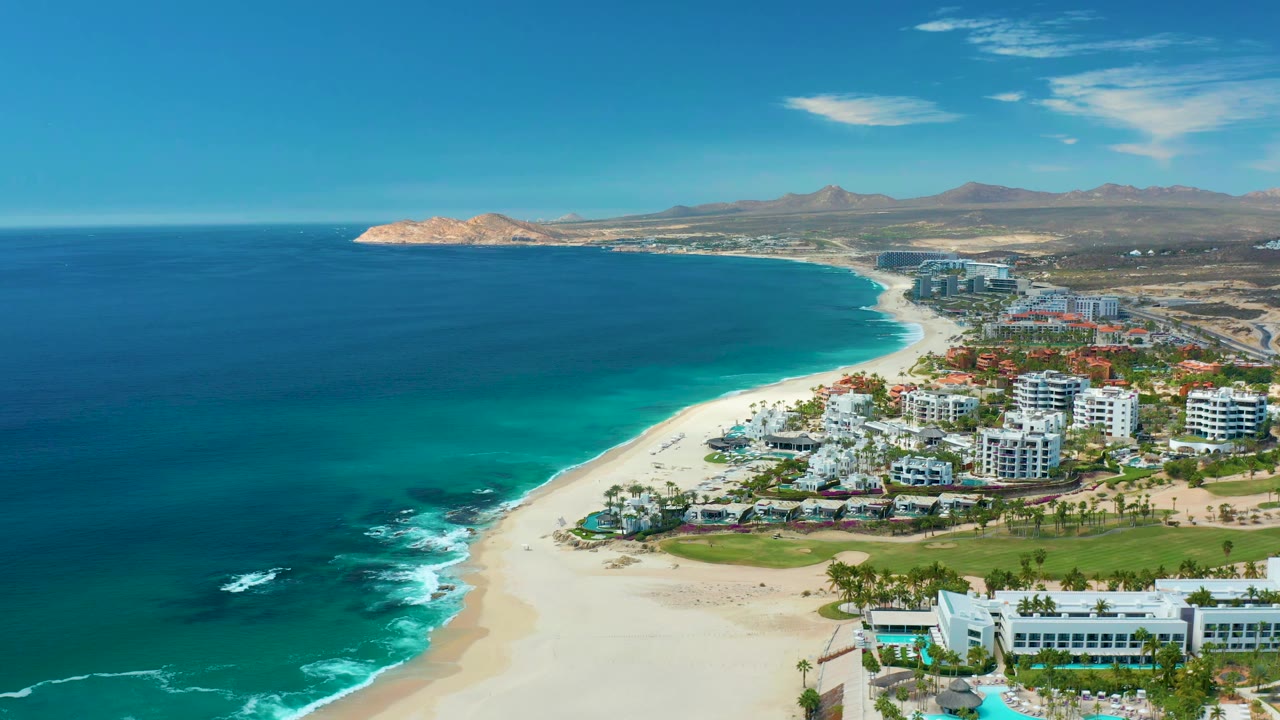 Aerial frame of Marquis Los Cabos, Mexico, showing the ocean occupying most of the left side, a bright sand shoreline through the center, coastal buildings on the right, and no visible people.