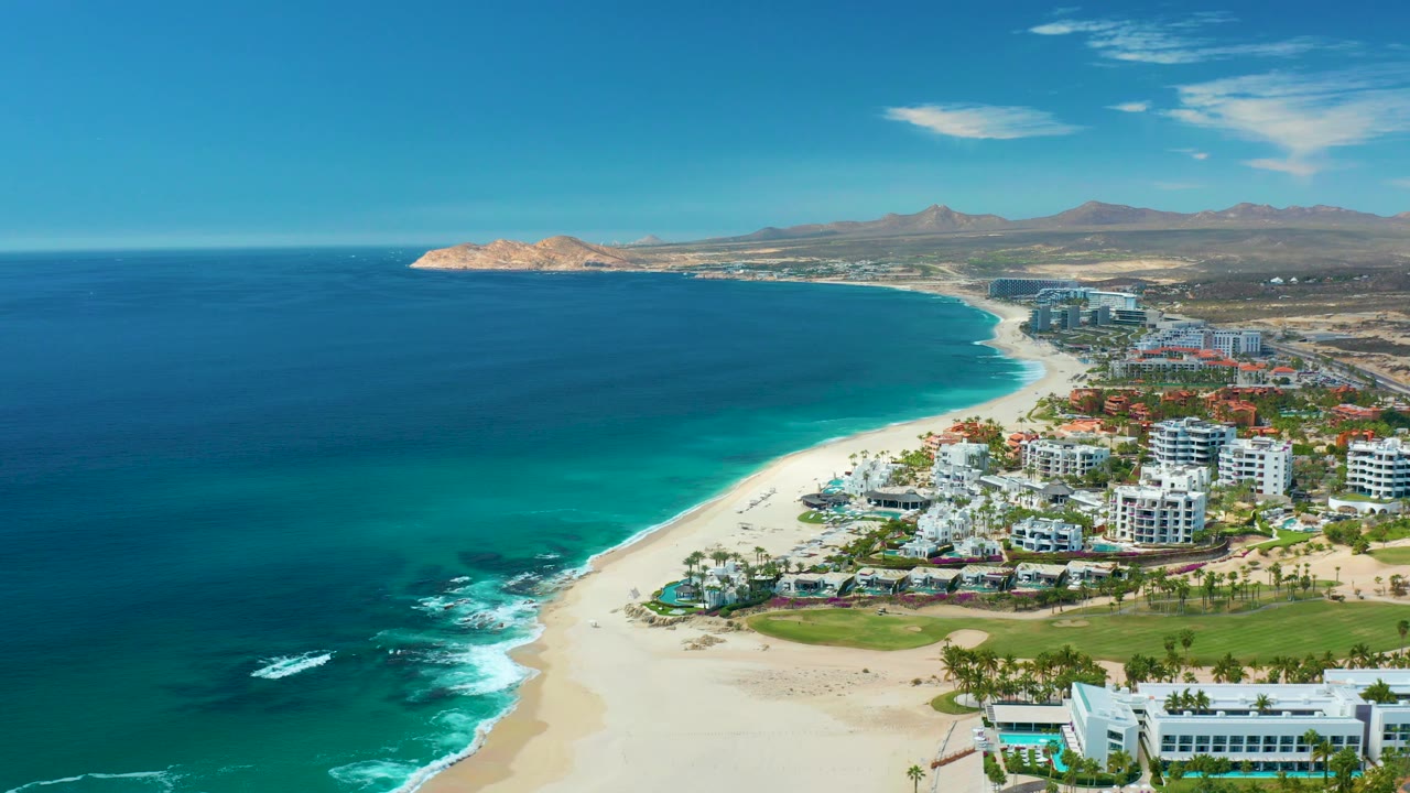 Mid-clip aerial frame over Marquis Los Cabos, Mexico, with more open water on the left, white surf touching the lower shoreline, resort blocks on the right, and no visible people.