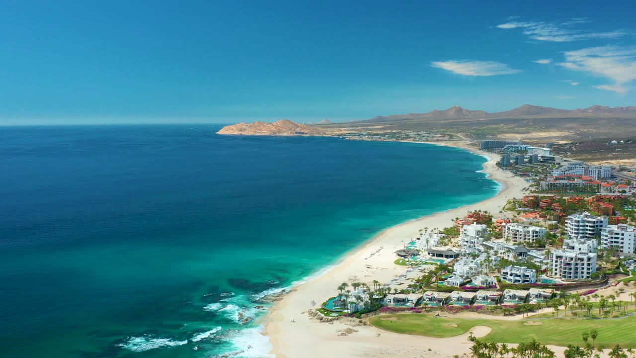 Wide aerial view of Marquis Los Cabos, Mexico, with deep blue ocean on the left, turquoise shallows, pale sand, beachfront buildings, and dry hills under bright sun.