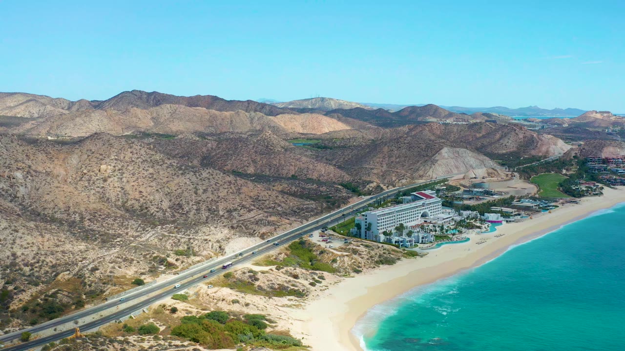 High oblique aerial view of highway traffic beside a sandy beach and turquoise water.