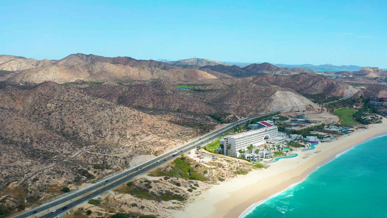 Aerial frame over Marquis Los Cabos, Mexico, showing cars on the highway, the hotel complex closer to center, bright sand and turquoise water to the right, and no visible people.