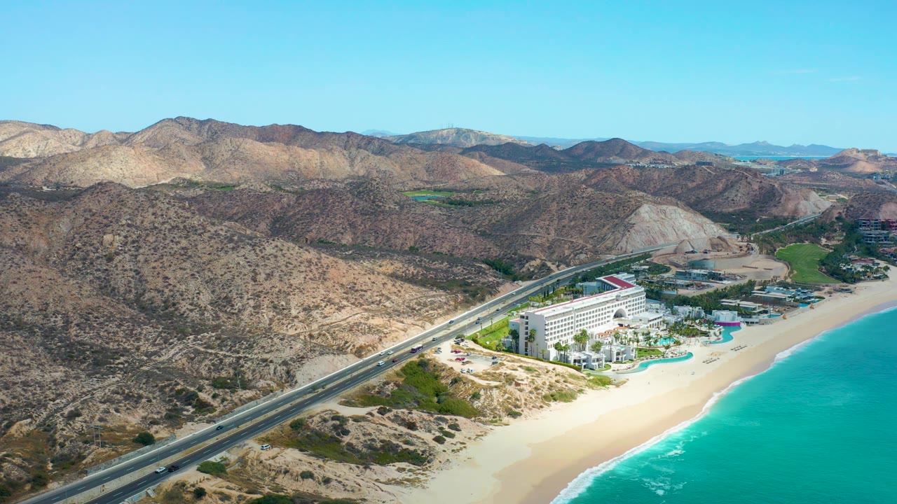Wide aerial view in Marquis Los Cabos, Mexico, with the highway angling past the hotel, pale sand meeting turquoise ocean, brown mountain ridges beyond, and no visible people.