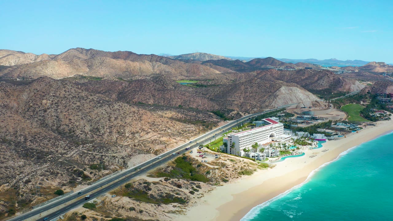 Aerial frame in Marquis Los Cabos, Mexico, with the beach and ocean filling more of the right edge, the hotel below the highway, layered desert hills in back, and no visible people.