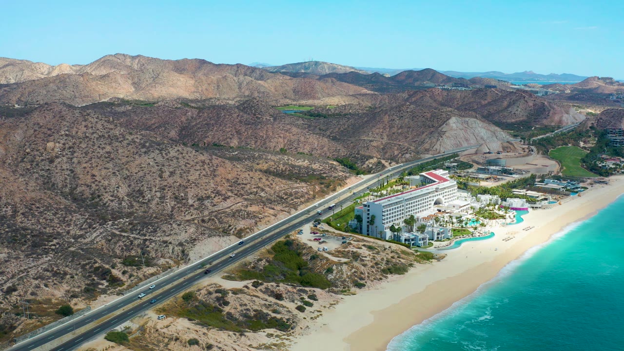 High aerial frame of Marquis Los Cabos with vehicles on the diagonal coastal highway, pale sand, bright turquoise water, beachfront buildings, dry hills, and no visible people.